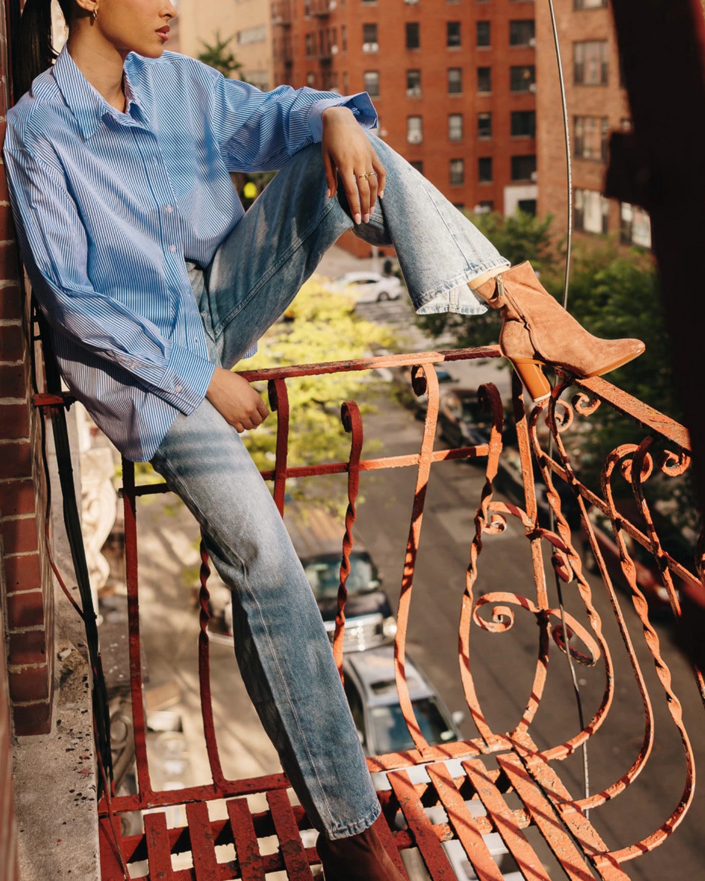 Person sitting on a balcony with a cityscape background in brown suede ankle boots