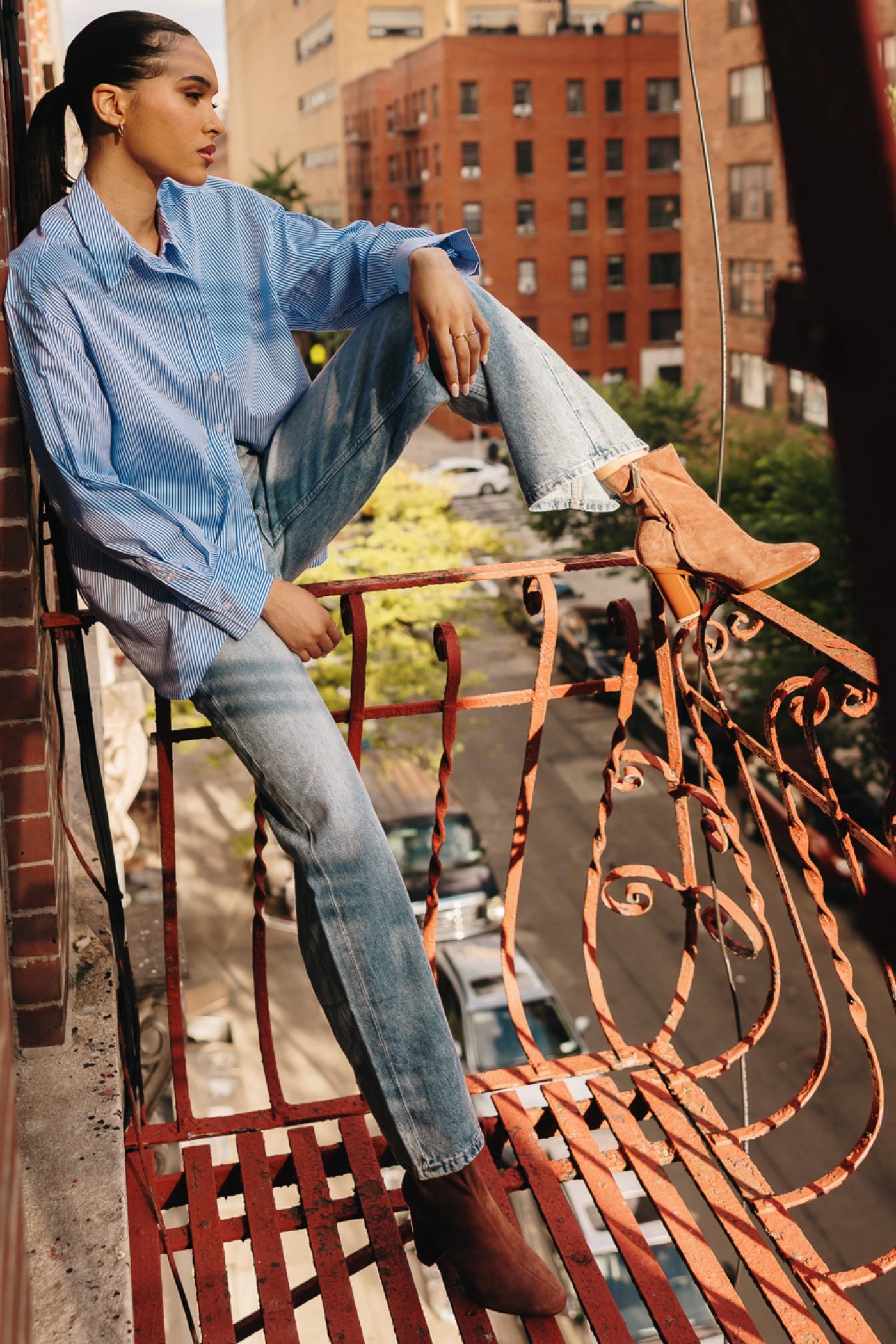 Person sitting on a balcony with a cityscape background in brown suede ankle boots