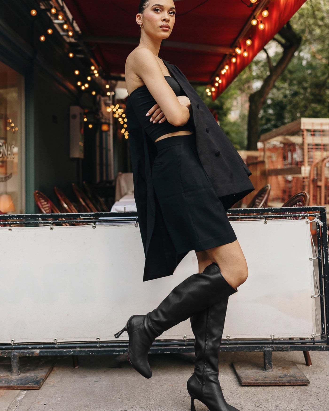 Woman in a black outfit with black knee-high boots standing in front of a restaurant with string lights.