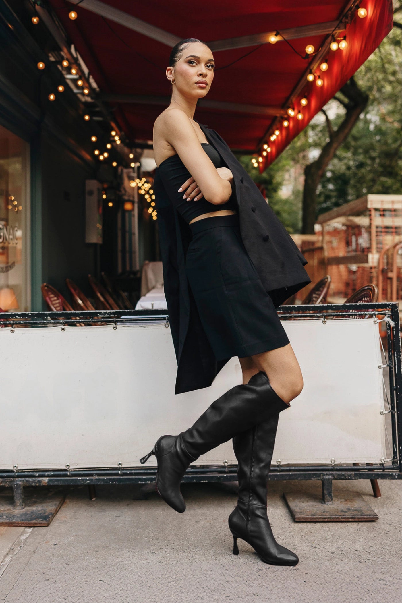 Woman in a black outfit with black knee-high boots standing in front of a restaurant with string lights.