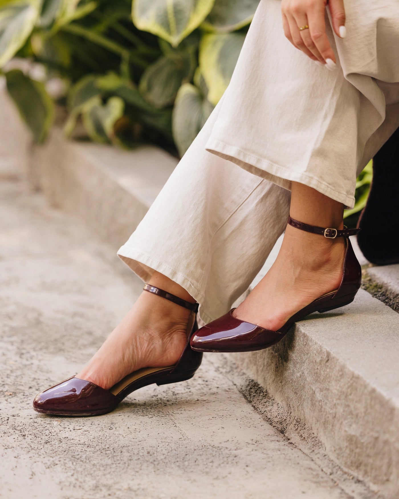 Person wearing burgundy closed toe flats and beige pants sitting on steps with greenery in the background