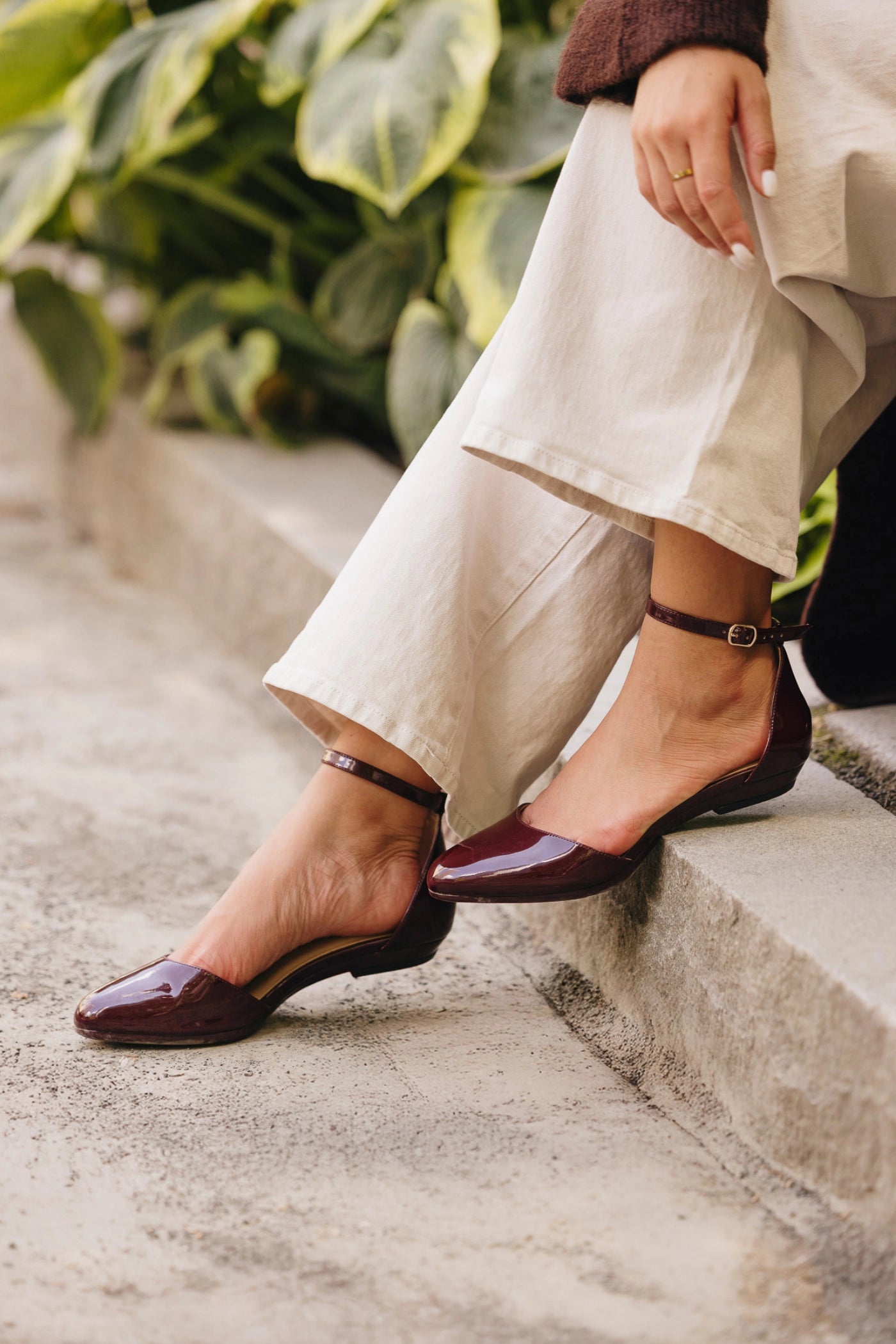 Person wearing burgundy closed toe flats and beige pants sitting on steps with greenery in the background