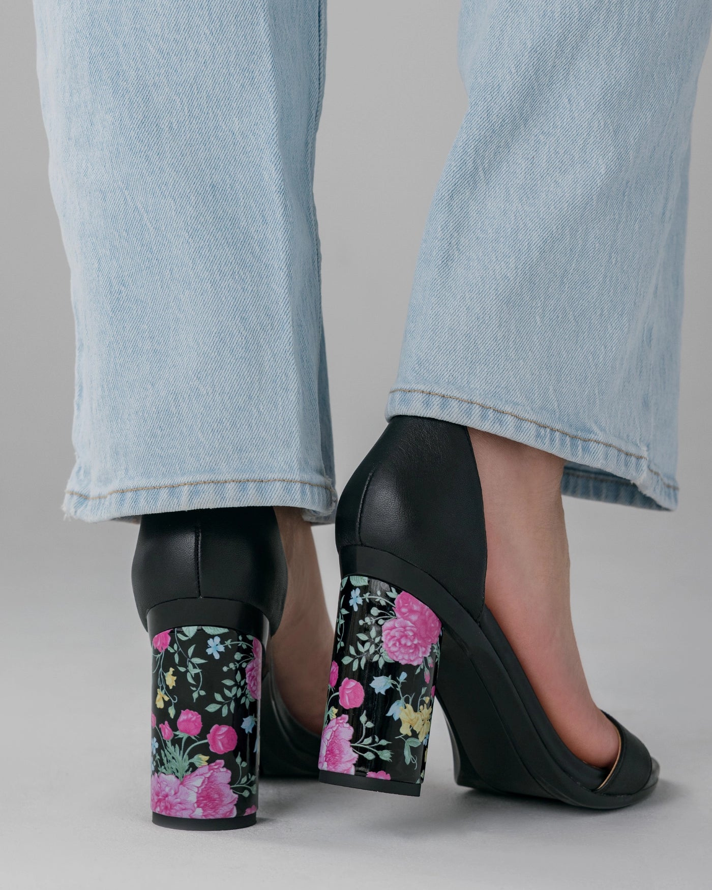 Black high-heeled shoes with floral design worn with light blue jeans on a neutral background