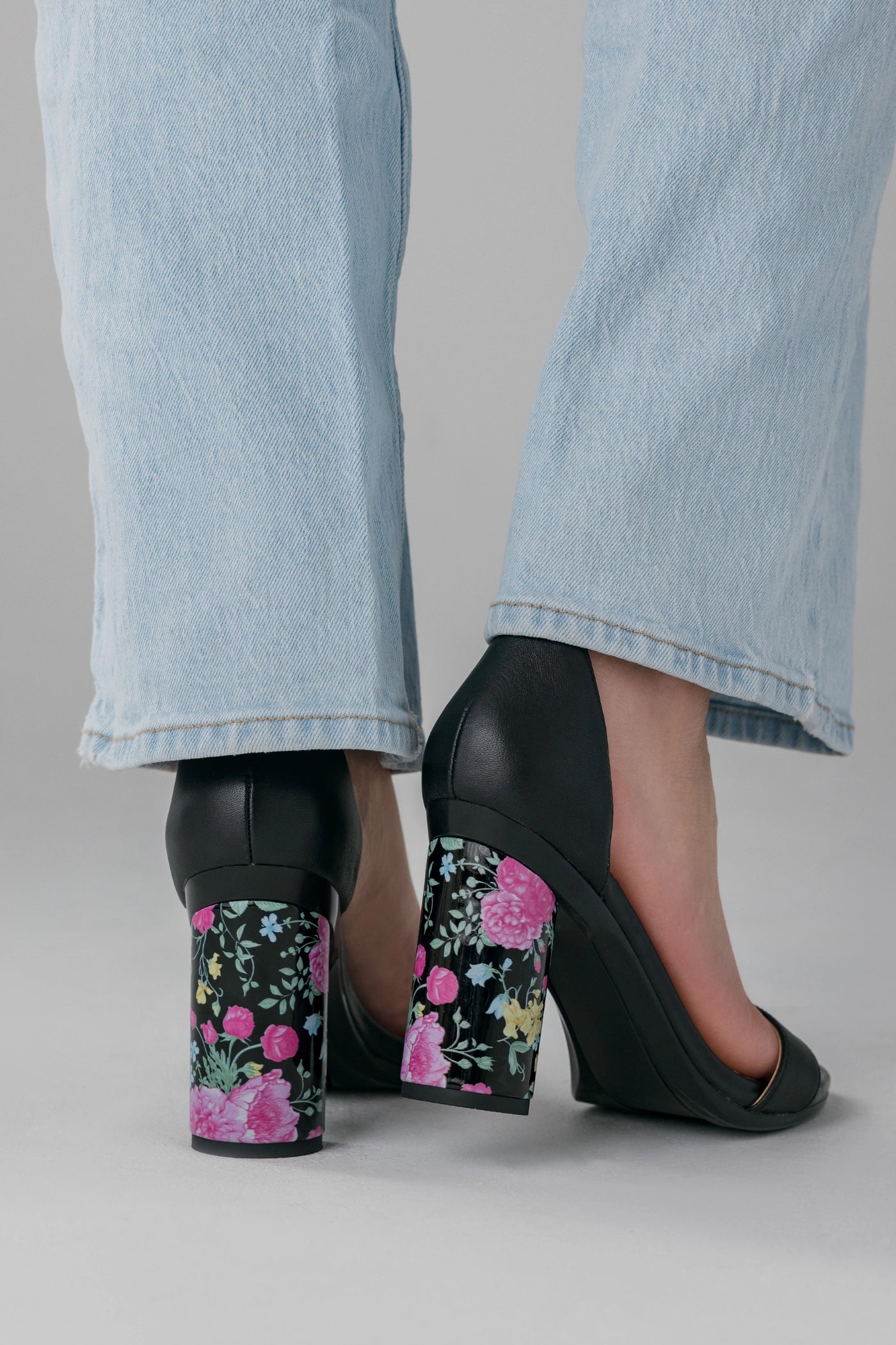 Black high-heeled shoes with floral design worn with light blue jeans on a neutral background