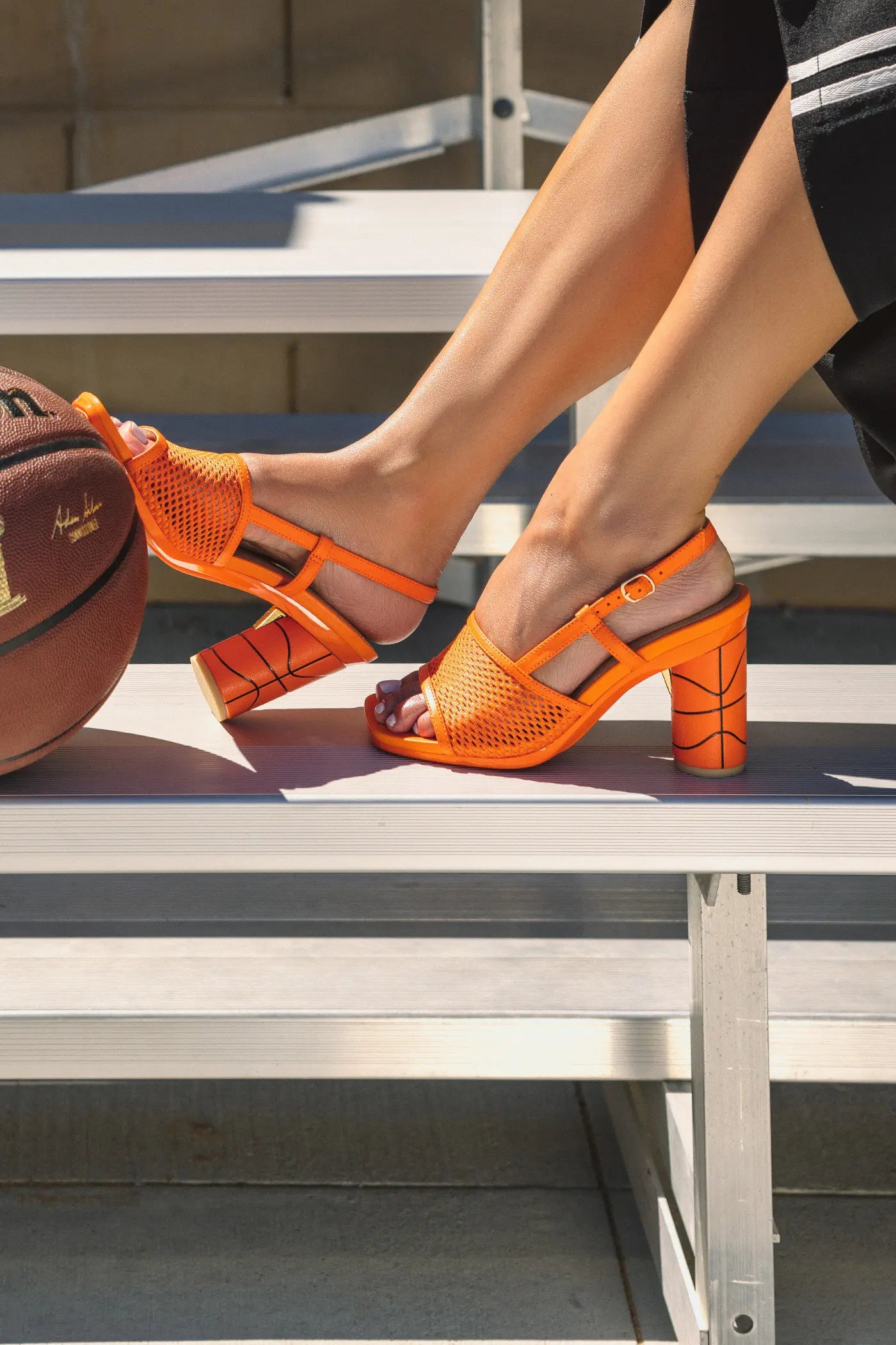 Orange sandals with basketball heel worn by a person sitting on a metal bench.